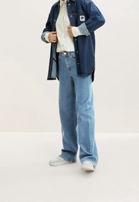 Giacca di jeans blu scuro con jeans blu chiaro. La giacca presenta una tasca con patta sul petto; entrambi i capi sono di taglio ampio. Le sneakers bianche completano il look.