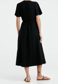 Robe noire jusqu'aux genoux avec manches courtes, tissu texturé, taille cintrée avec détail à nouer, et une silhouette ample et fluide. Portée avec des sandales.
