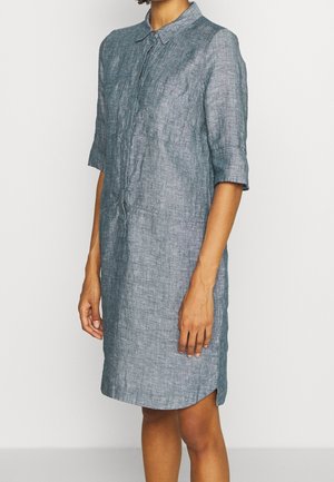 Donna che indossa un vestito grigio di lino, corto al ginocchio, con maniche corte, bottoni e colletto, su uno sfondo bianco uniforme.