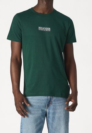 Homme portant un t-shirt vert foncé à manches courtes avec le texte "HILFIGER COLUMBUS AVE. NEW YORK" sur la poitrine et un jean bleu clair.