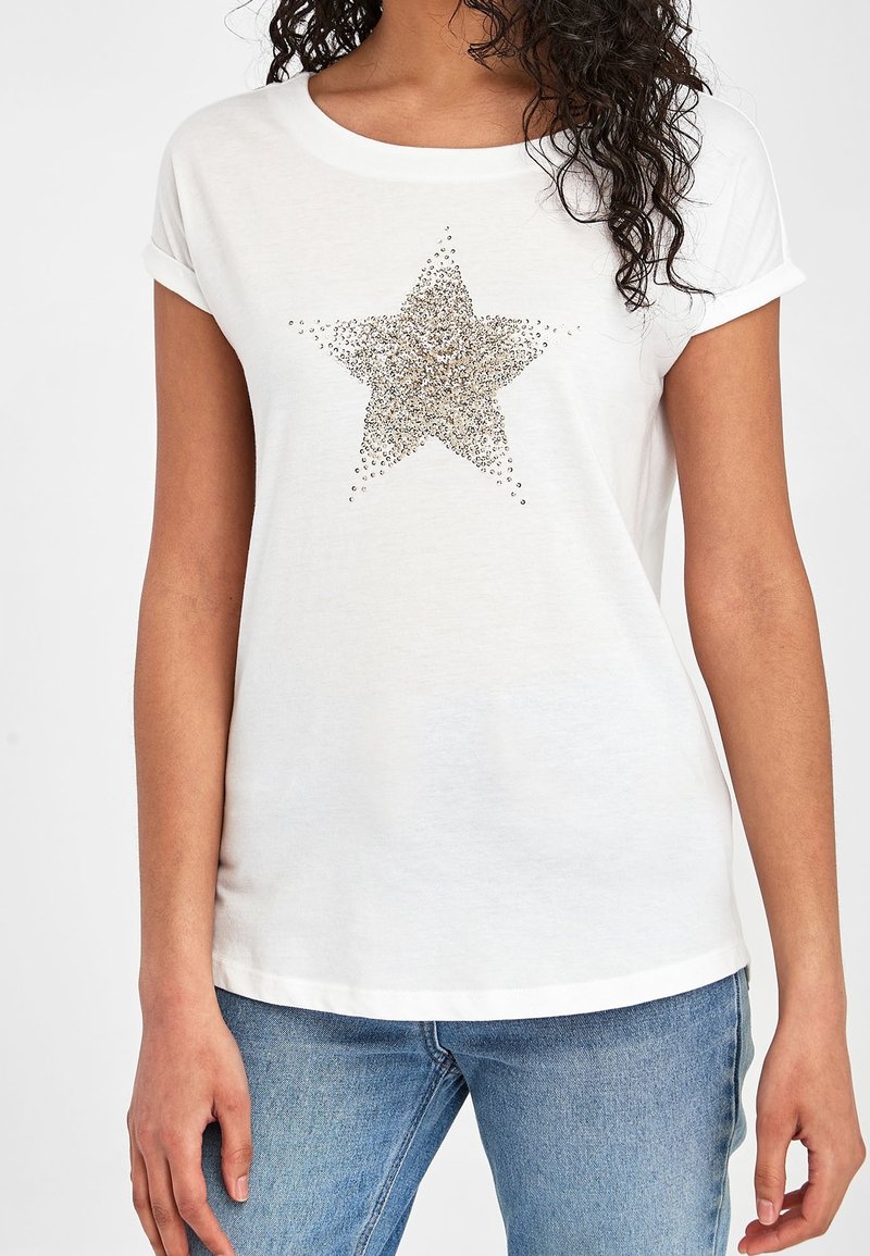 T-shirt blanc à manches courtes présentant un grand motif d'étoile dorée scintillante composé de petits strass au centre, avec des ourlets de manches roulés.