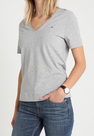 Kvinde iført en lysegrå kortærmet V-hals T-shirt, kombineret med blå jeans, et armbåndsur og hånden i lommen.