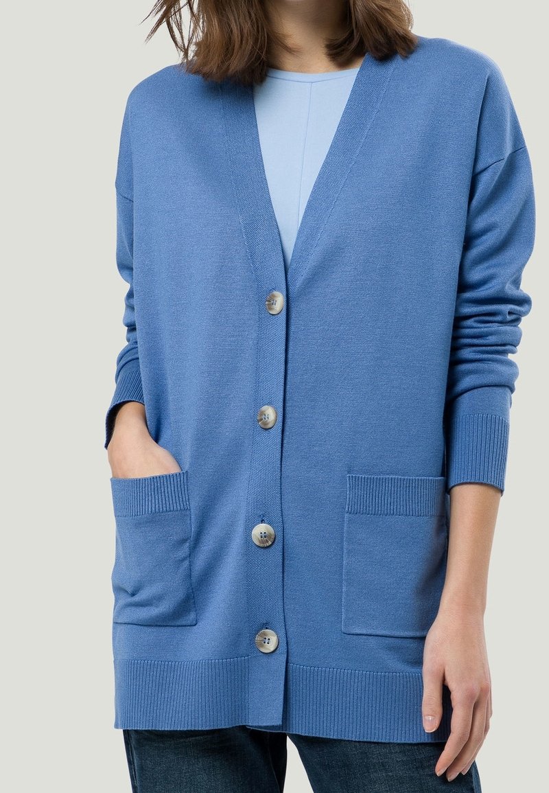 Modrý kardigan vyrobený z měkké tkaniny, s výstřihem do V, pěti knoflíky, dvěma předními kapsami a žebrovanými manžetami a lemem.