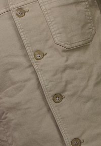 Beige bomullskjorta med fem synliga knappar, en bröstficka och sydda detaljer längs sömmarna. Mjuk textur med en matt yta.