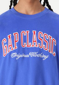 Camiseta de algodón azul con el texto en negrita rojo y blanco "GAP CLASSIC", con "Ropa Original" en cursiva debajo. Cuello redondo y mangas cortas.