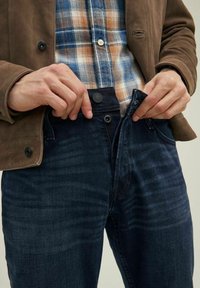 Jeans in denim scuro con una finitura testurizzata, abbinati a una camicia a quadri blu, arancione e bianco, e una giacca in suede marrone.
