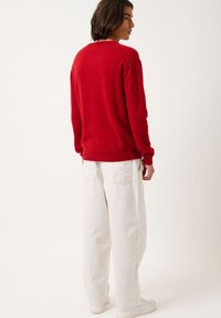 Maglione rosso a maglia con maniche lunghe, scollo rotondo e polsini a coste. Abbinato a pantaloni bianchi larghi e sneakers bianche. Vista da dietro.