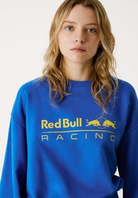 Plava dukserica s odvažnim žutim logotipom "Red Bull Racing" uz grafičke akcentne detalje, okruglim izrezom i ležernim krojem.