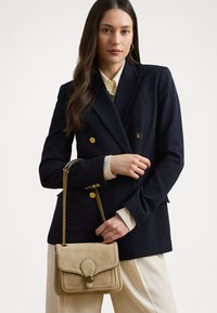 Borsa in suede beige con pattina, dettagli dorati e tracolla a catena. Il modello indossa un blazer blu navy e una camicia crema, evidenziando un design moderno e strutturato.