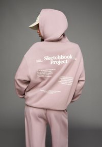 Persoon in een lichtroze hoodie en broek, met de tekst "Sketchbook Project" en andere zinnen op de achterkant van de hoodie, staand tegen een grijze achtergrond.