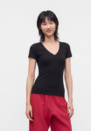 Jeune femme aux cheveux noirs courts, vêtue d'un t-shirt noir à col en V et d'un pantalon rouge ample, debout devant un fond clair uni.