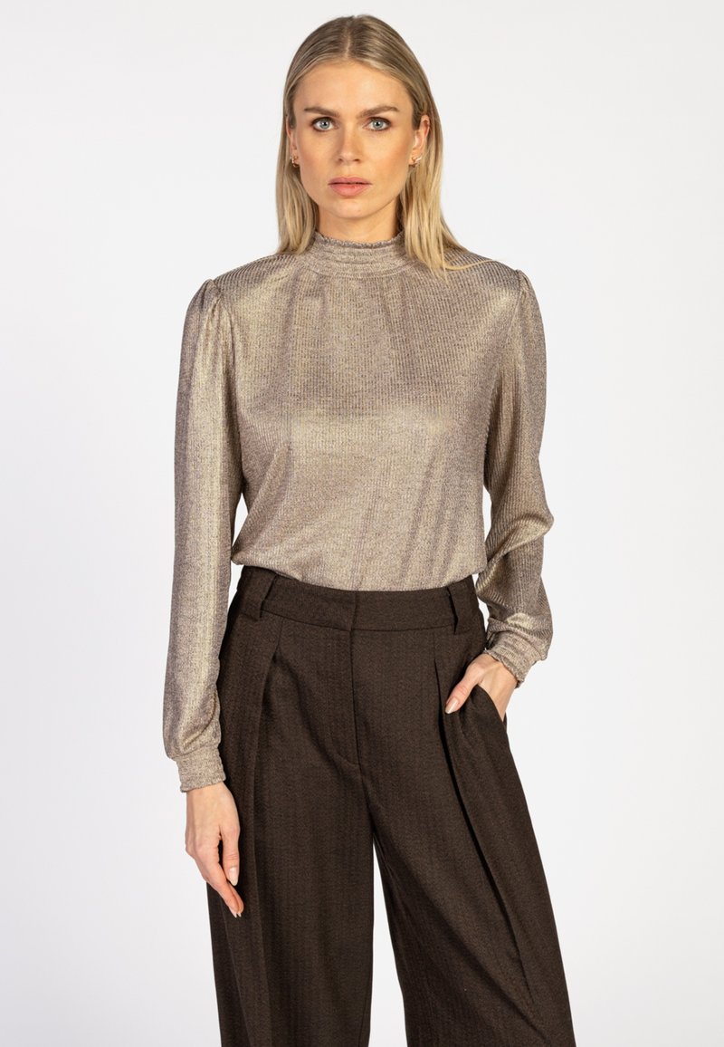 Vrouw met blond haar, gekleed in een metallic lange mouw top en een donkerbruin hoge taille wijde broek, staande tegen een effen achtergrond.