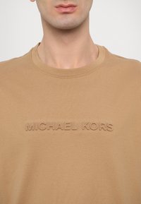 Tan cotton t-shirt featuring a round neckline and embossed "MICHAEL KORS" text across the chest. Soft texture, casual fit.