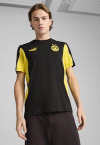 Černé a žluté tričko s krátkým rukávem se znakem Puma a symbolem BVB. Má kulatý výstřih, kontrastní barevné bloky a hladkou texturu.