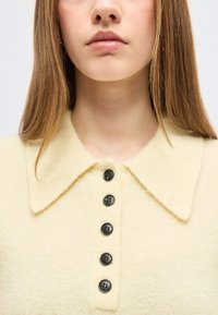 Νεαρή γυναίκα με ίσια ανοιχτοκάστανα μαλλιά φοράει ένα παλ ανοιχτό κίτρινο πλεκτό πουλόβερ με μεγάλο γιακά και πέντε μαύρα κουμπιά στο εμπρός μέρος.