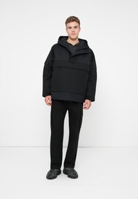 Casaco preto com capuz e design minimalista, apresentando um bolso frontal e cordões ajustáveis, combinado com calças e sapatos pretos.