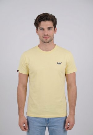 Joven con cabello corto y castaño, vistiendo una camiseta de manga corta color amarillo claro y jeans azul claro, de pie contra un fondo gris liso.