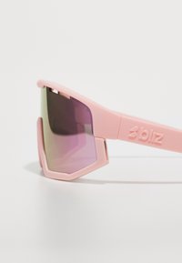 Ochelari de soare sport roz, cu lentile unghiulare, supradimensionate, care se tranziționează de la verde la purpuriu, având un finisaj mat și un logo embossat pe lateral.