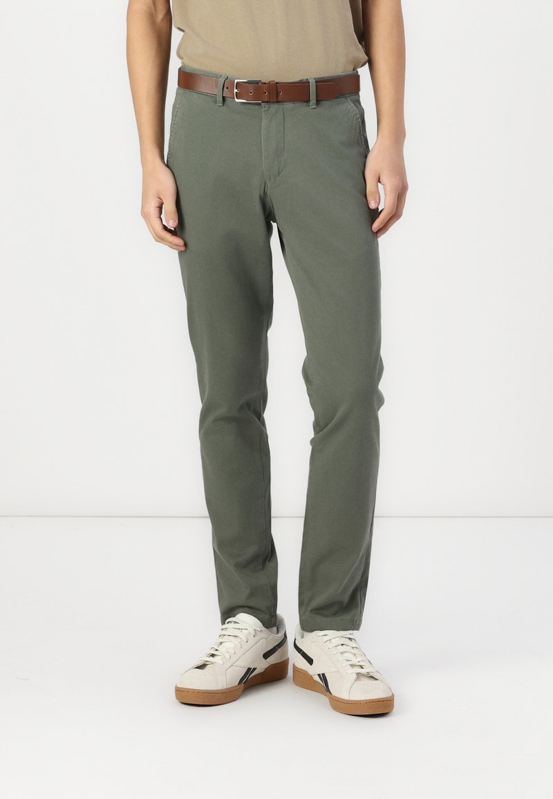 Pantalons en coton verts avec une coupe droite, dotés de poches latérales et d'une ceinture en cuir marron, associés à des chaussures blanches avec des accents noirs.