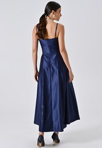 Marineblauwe satijnen jurk met een aansluitende bustier en een uitlopende rok. Kenmerkt zich door dunne verstelbare banden en een gladde textuur, gecombineerd met donkere hakken.