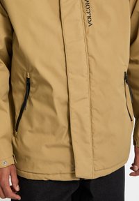 Tan zip-up jakke med en glat stofstruktur, der har to sidelommer med sorte lynlåse og et logo-print foran.