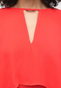 Moteris dėvi raudoną palaidinę su giliu V formos iškirpte, prisegtą metaliniu sagčiu ties raktikauliu.