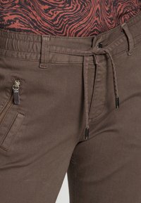Brown pants with an elastic waistband and drawstring; features a zipper pocket on the side, styled against a patterned top.