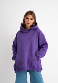 Felpa oversized viola con una grande tasca frontale e cappuccio con cordino, abbinata a jeans azzurri chiari. Texture liscia e design casual.