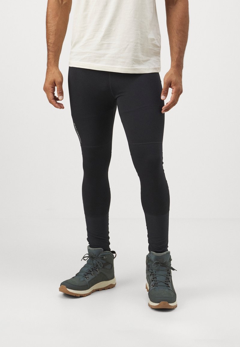 Homme portant des leggings noirs ajustés et des bottes de randonnée vert foncé, debout devant un fond blanc uni.