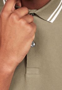 Polo khaki avec un col classique, featuring des rayures blanches, trois boutons, et un tissu doux et texturé en gros plan sur le col et la main.