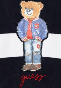 Marineblaue Stoff mit weißen Streifen zeigt einen Bären in einer blauen Collegejacke und Jeans, versehen mit rotem Text "guess." Gestickte Elemente.