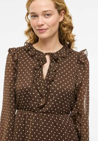 Brun prikkete kjole med rysjekrage og ermer, har en åpning ved halsen og en flytende silhuett. Laget av lett stoff.