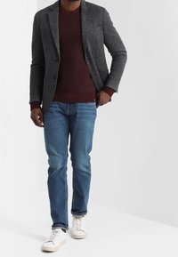 Grauer Blazer über einem burgunderfarbenen strukturierten Pullover, kombiniert mit blauen Jeans und weißen Sneakers. Der Blazer hat eine Reverskragen und eine Brusttasche.