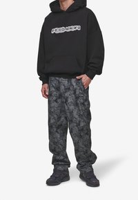 Sweat à capuche noir avec un logo, associé à un pantalon ample gris à motif camouflage et des baskets foncées. Comprend une poche avant et des poignets côtelés.