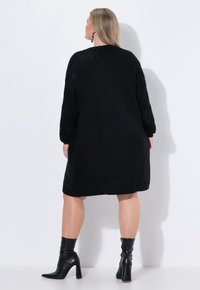 Černé pletené oversized šaty s dlouhými rukávy a kulatým výstřihem, se strukturovaným vzorem a žebrovaným lemem, doplněné černými botami na podpatku.