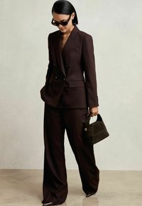 Blazer croisé marron foncé avec un pantalon large assorti, accompagné d'un petit sac à main en daim foncé, présentant un design structuré.