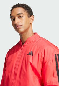 Red bomber jacket with a front zipper, black logo on chest, and black stripes along sleeves. Made of smooth, lightweight material.