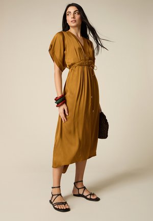 Donna che indossa un abito midi color senape con vita stretta, sandali neri con borchie, braccialetti colorati e tiene in mano una borsa scura con texture.