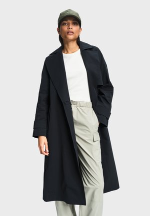 Žena koja nosi dugi crni baloner, bijelu košulju, svijetlo bež cargo hlače i zelenu kapu na jednobojnoj sivoj pozadini.