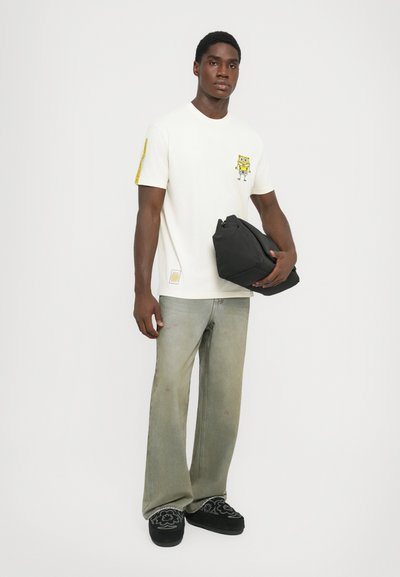 Vit t-shirt med en gul SpongeBob SquarePants-grafik, parat med ljusgröna utsvängda jeans och svarta slip-on skor. Svart väska i handen.