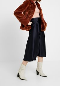 Femme portant une veste en fausse fourrure couleur rouille, un haut côtelé rose, une jupe midi en satin noir et des bottes cowboy beiges à talons, posant devant un fond blanc.