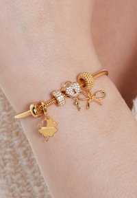 Gouden armband met bedels in de vorm van een hart, strik, sleutel en klavertje vier, afgewisseld met gestructureerde en gladde kralen, met sprankelende details.