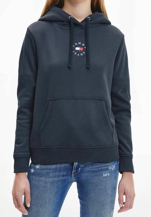 Vrouw draagt een donkerblauwe hoodie met een voorzak en Tommy Jeans-logo, gecombineerd met blauwe distressed jeans.