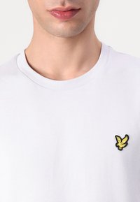 Vit bomulls t-shirt med rund halsringning och en liten broderad fågel-logotyp i gult och svart på vänster bröst.