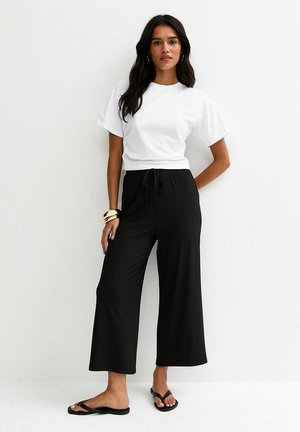 Vrouw staat met hand op heup, draagt een wit T-shirt met korte mouwen, zwarte wijde broek met trekkoord, zwarte sandalen en gouden armbanden.