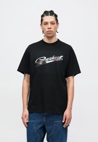 Czarny bawełniany t-shirt z błyszczącym, srebrnym logo "Carhartt" w stylu pisma odręcznego, krótkimi rękawami i luźnym krojem, noszony z niebieskimi dżinsami.