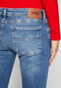 Vaqueros de mezclilla azul con una altura media, que presentan una etiqueta de cuero con la inscripción "TOMMY JEANS" y un pequeño logo en el bolsillo trasero. Acabado desgastado y con textura.