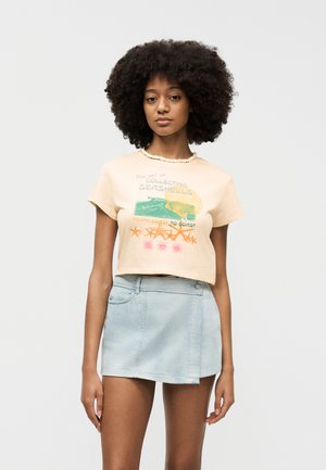 Νεαρή γυναίκα με σγουρά μαλλιά φοράει μπεζ γραφικό crop top και φωτεινή μπλε μίνι φούστα από τζιν, μπροστά σε απλό λευκό φόντο.