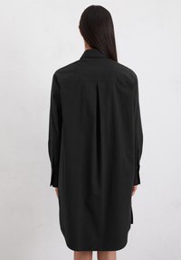 Zwarte jurk met kraag, lange mouwen en een achterpleat. Gemaakt van soepel materiaal, het heeft knoopmanchetten en zijsplitten.