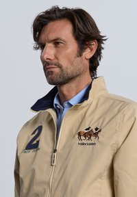 Chaqueta beige de tejido suave, con cuello azul, diseño de jugador de polo bordado y el número "2" en navy.
